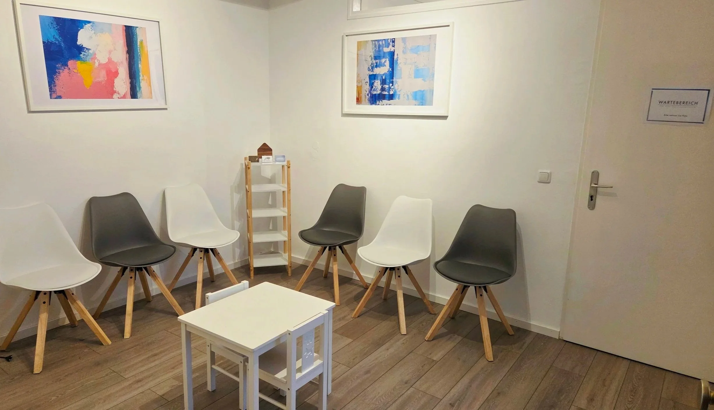 Warteraum mit sechs Stühlen in Schwarz und Weiß, zwei weißen kleinen Tischen, Wanddekoration mit zwei bunten abstrakten Kunstwerken, Holzregal mit kleinen Gegenständen, Tür mit Schilder, Holzfußboden.