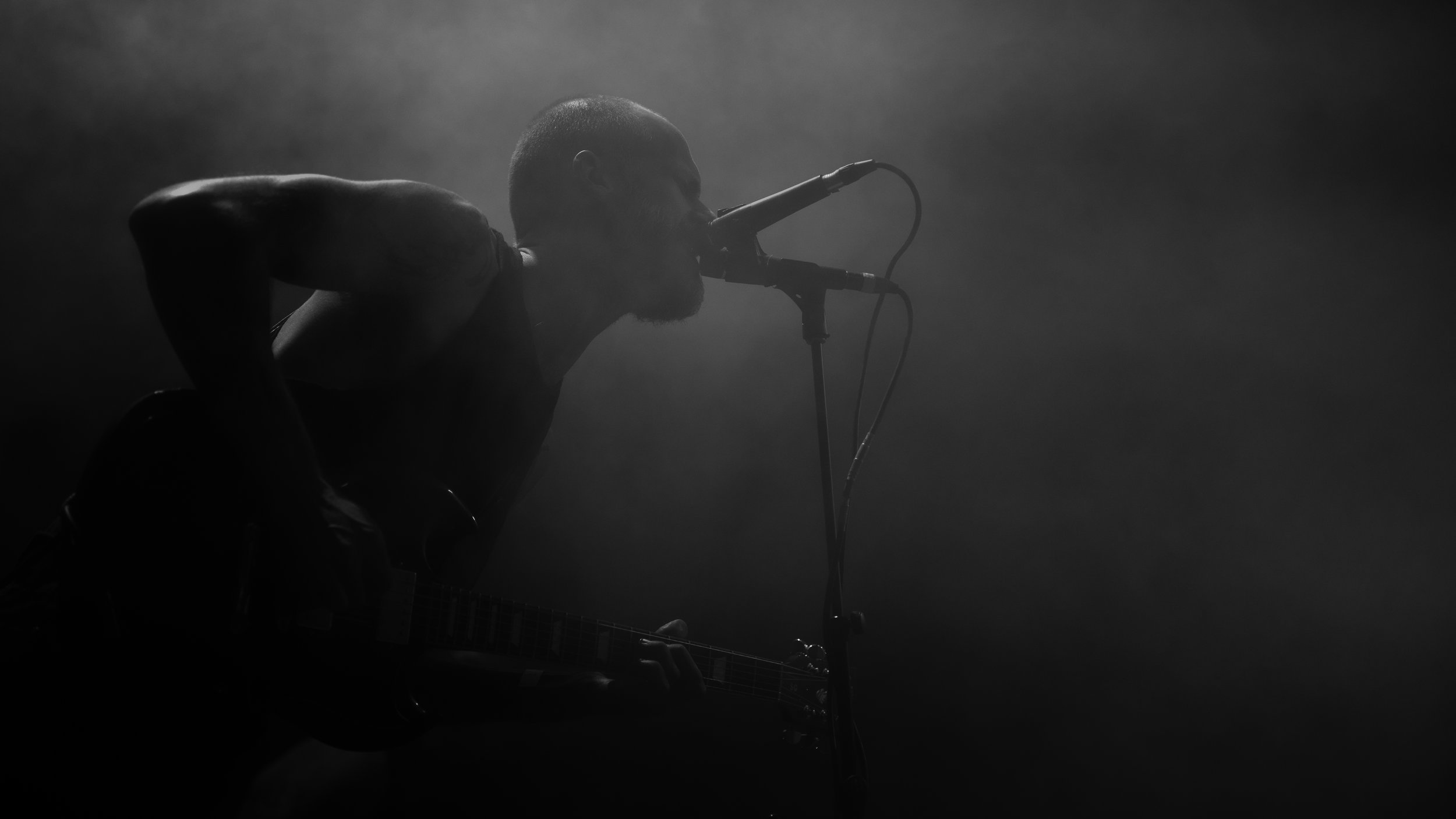 A black and white photo of a man singing into a microphone while playing guitar, with a plain dark background.