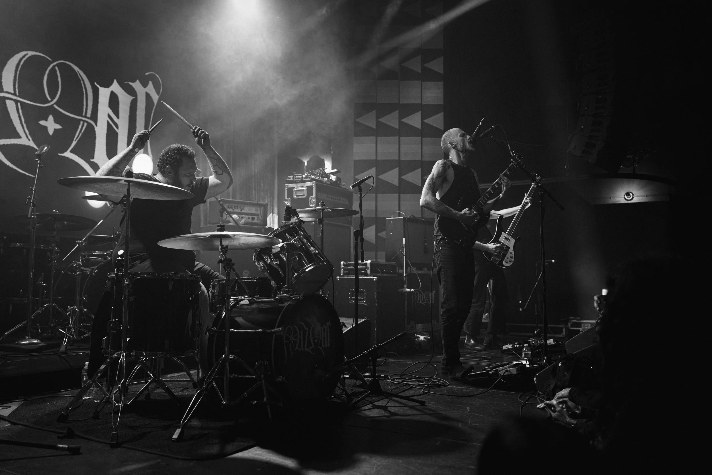 A black and white photo of two musicians performing on stage. One is playing drums and the other is singing and playing guitar. There are musical equipment and a backdrop with a design behind them.