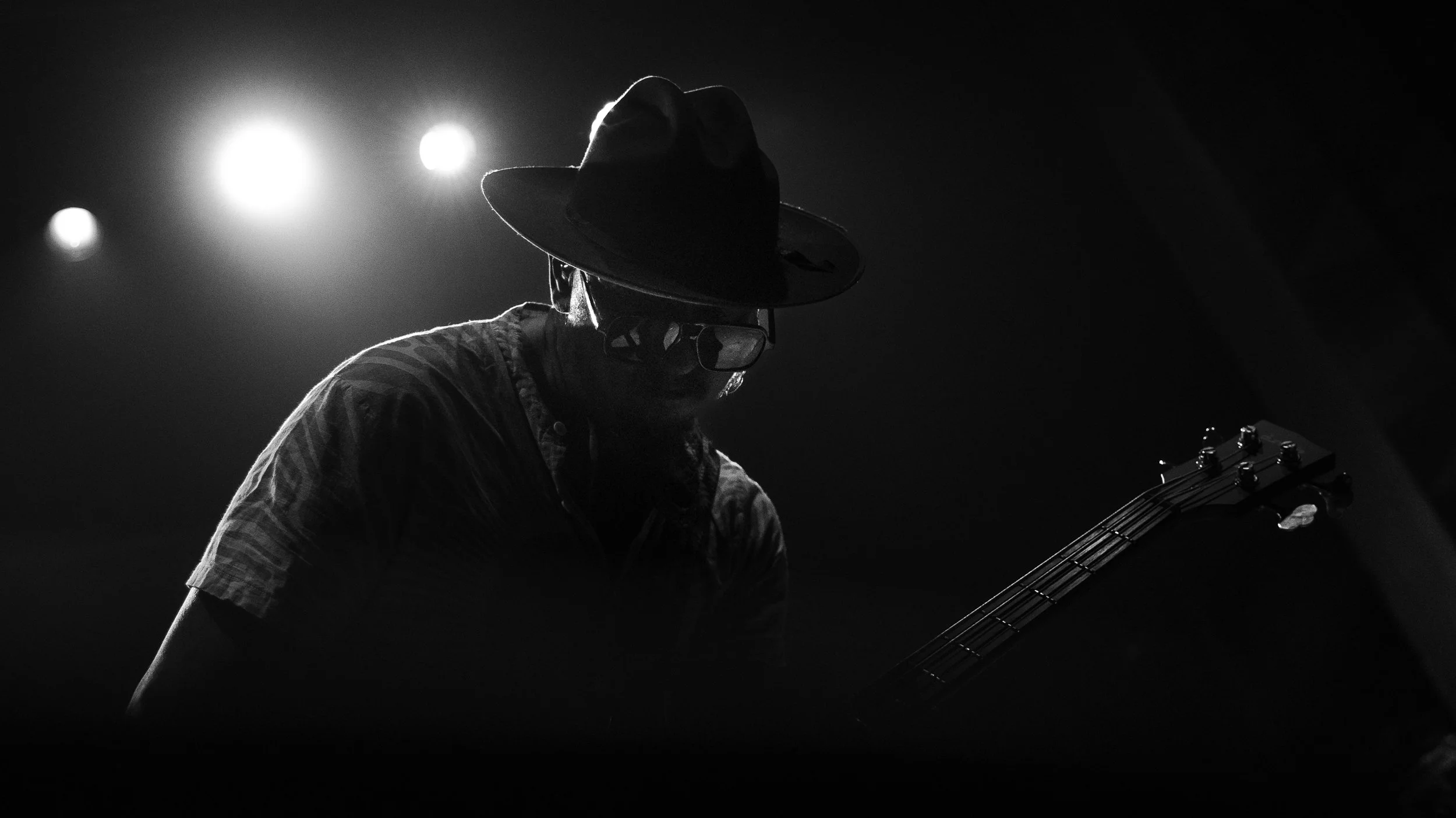 A person wearing a hat and glasses playing an electric guitar in a dark setting with bright lights in the background.