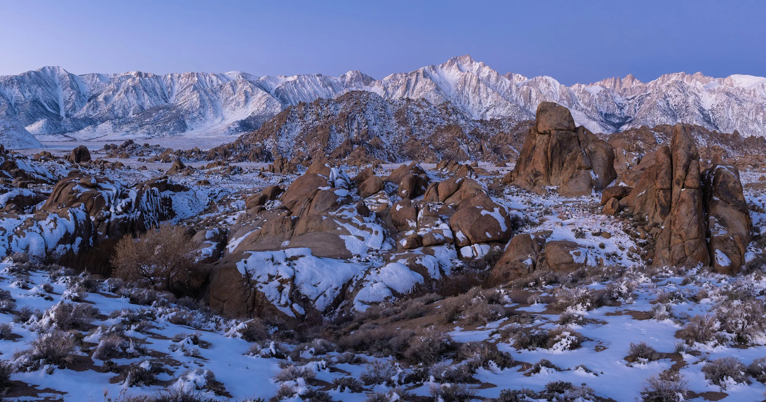 Snow-covered rocky desert with large boulders and mountains in the background under a clear blue sky.