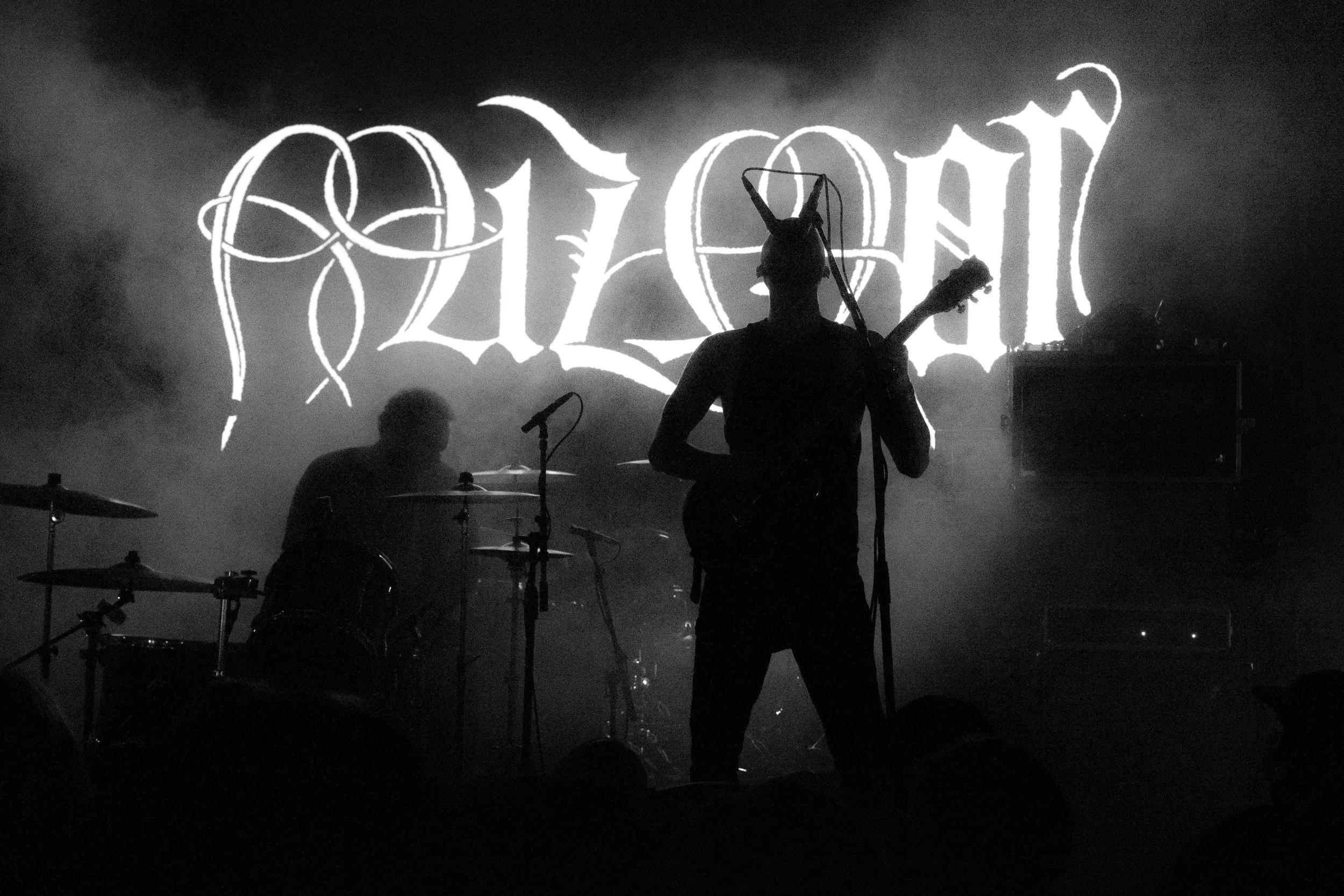 Silhouette of a musician with a guitar and a person in the background with a drum set on stage at a concert, with a large illuminated sign behind them that reads 'Darkthrone' in gothic style letters.