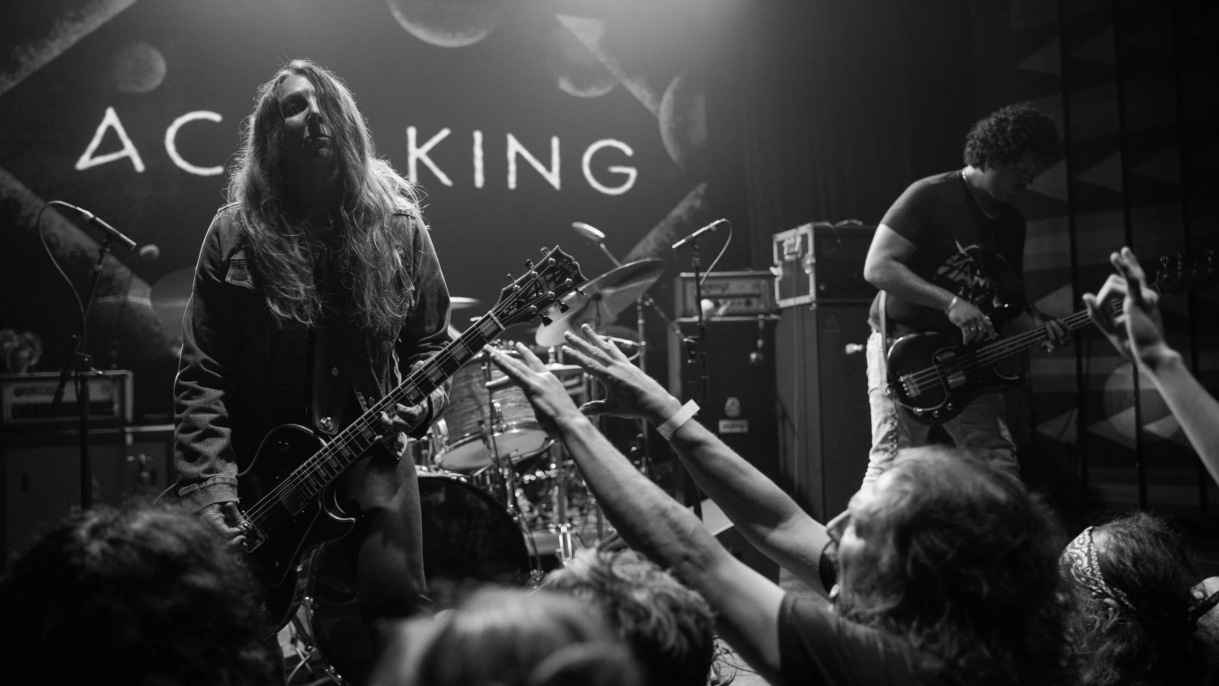 Black and white photo of a live music performance showing a guitarist and a bassist on stage, with audience members reaching out toward them, and the word 'ACKING' visible in the background.
