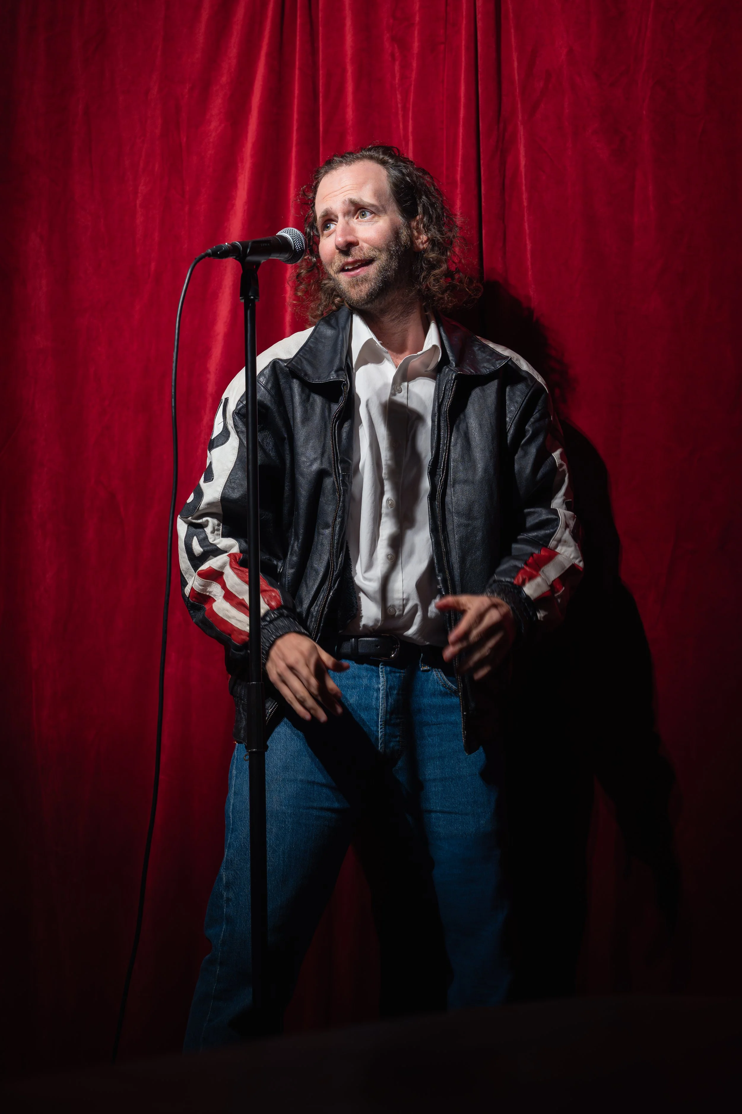 A man with curly brown hair and a beard stands on stage in front of a red curtain, wearing a leather and fabric jacket and blue jeans, speaking into a microphone.