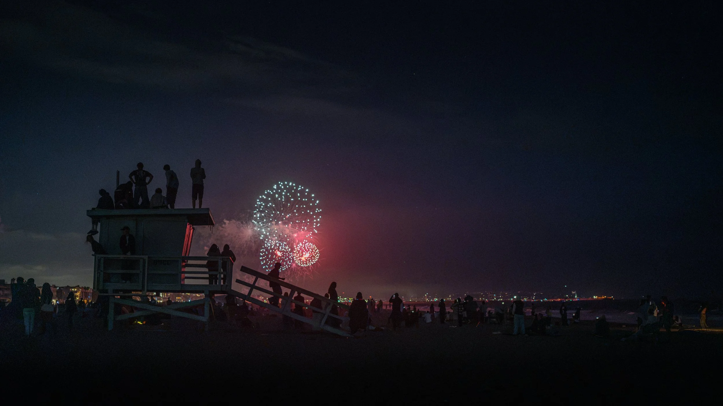 Silhouetted people on a pier watching fireworks in a dark night sky.