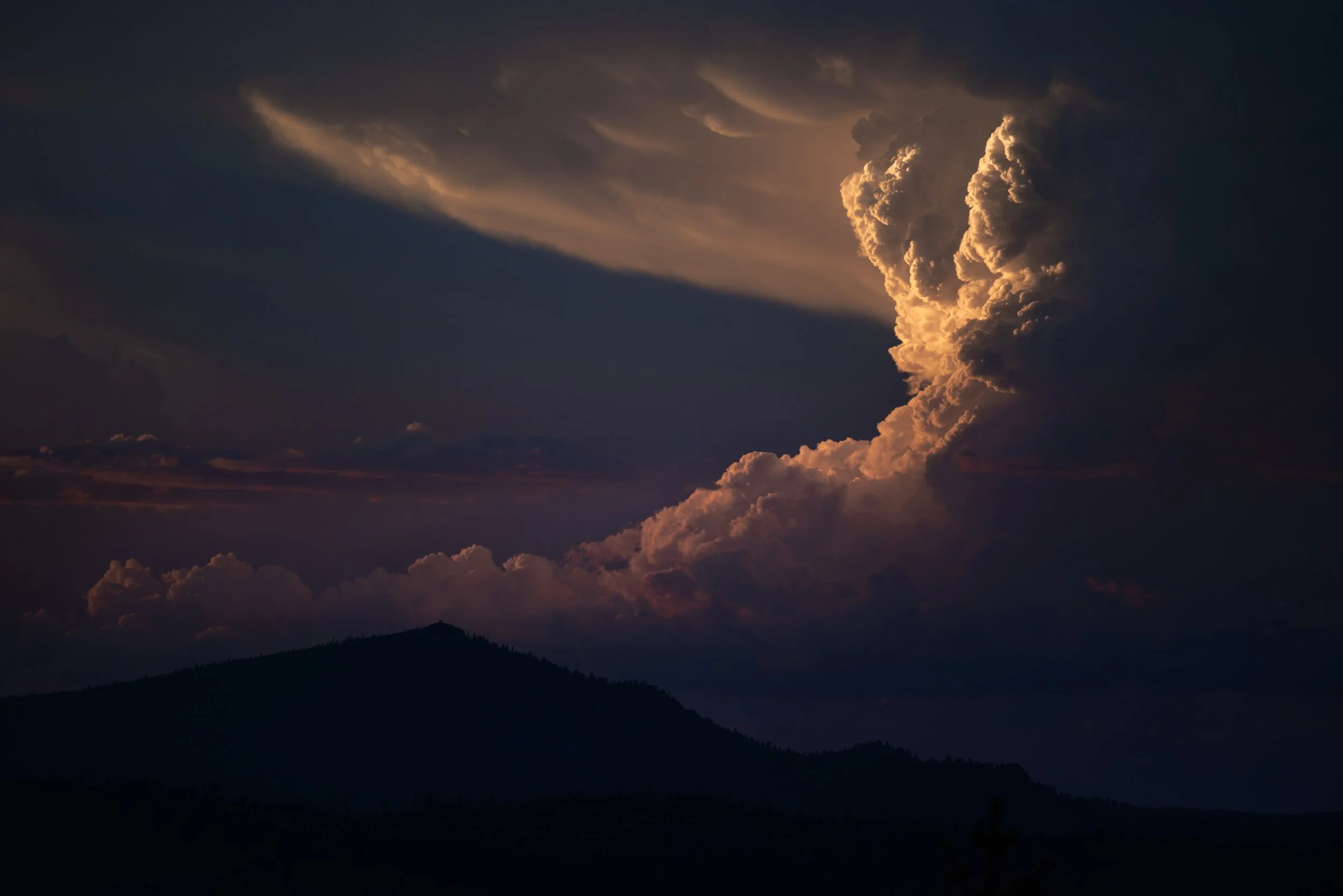 A dark sky with clouds illuminated by the setting or rising sun, casting a warm glow on the cloud formations. silhouette of mountains at the bottom.