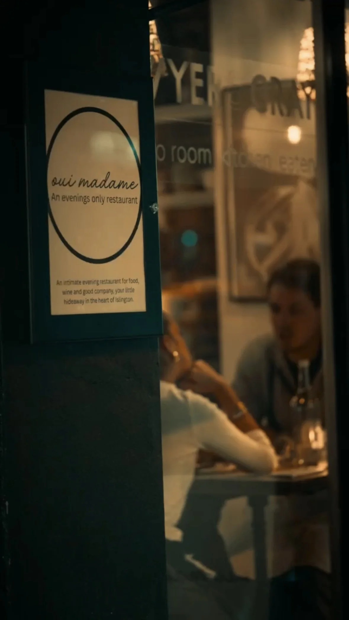 Nighttime scene of a restaurant named 'oui madame,' with visible signage describing it as an evening-only restaurant. Inside, two women are sitting at a table, engaged in conversation.