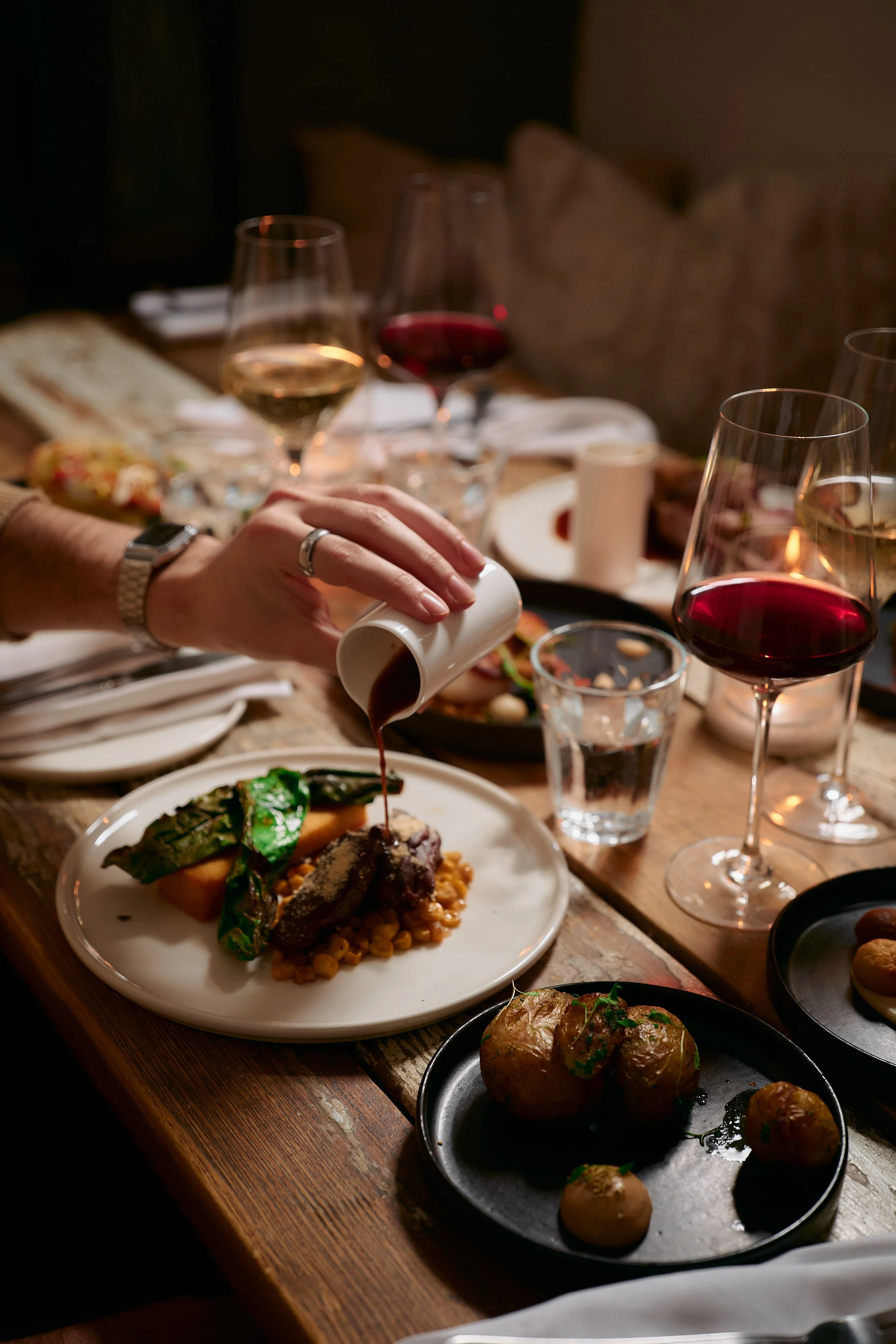 A dinner table with plates of food and glasses of red and white wine, where a person is pouring gravy on a meat dish.