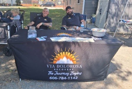 Two men sitting behind a table with a black tablecloth that has a mountain and sunrise logo and the text 'VIA DOLOROSA RECOVERY,' with contact information, outdoors in a residential area.