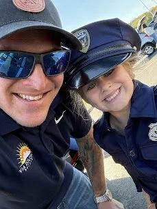 A man and a young girl smiling for a selfie outdoors, both wearing police hats and sunglasses.