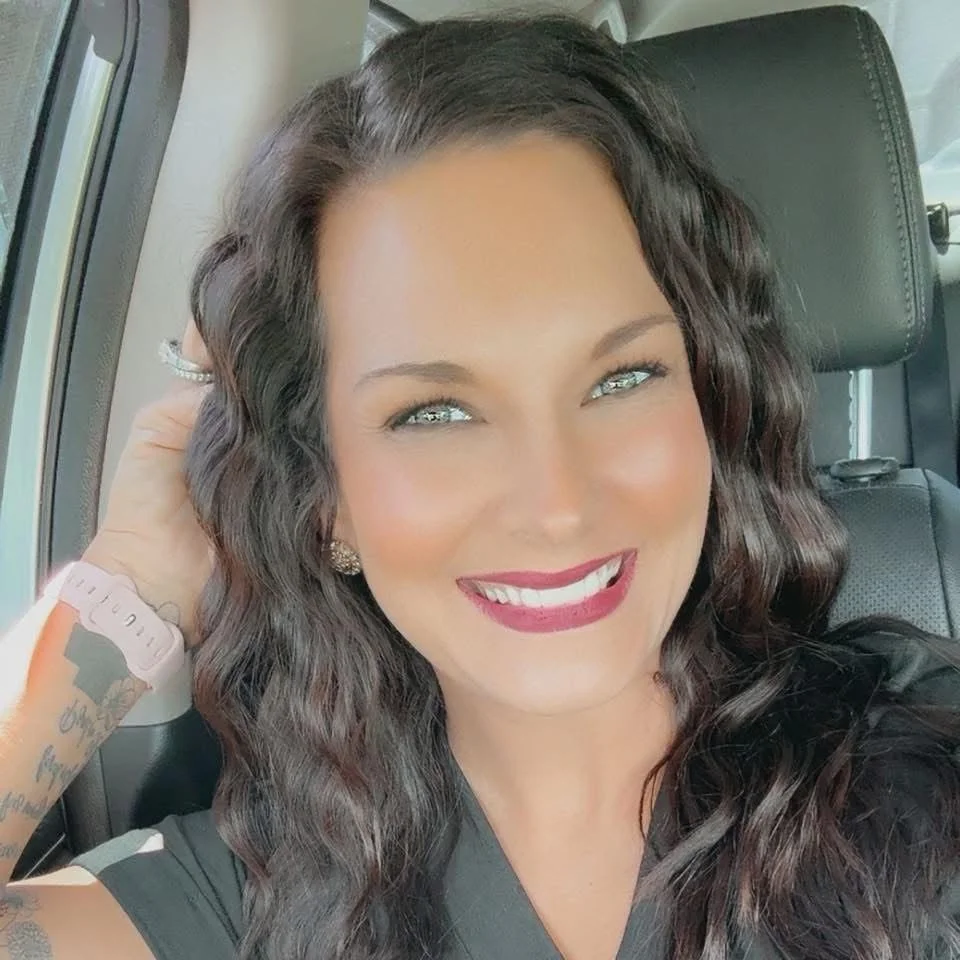 A woman with wavy dark hair and light-colored eyes smiling inside a vehicle.