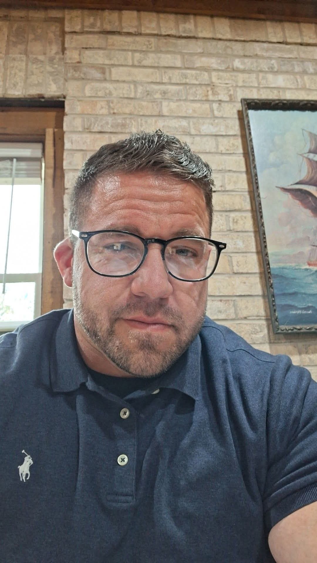 A man with short hair and glasses taking a selfie indoors, with a brick wall and a painting of a sailing ship in the background.