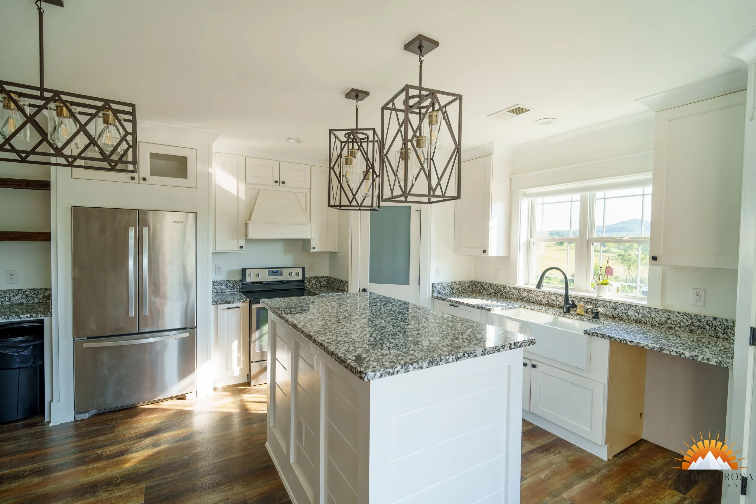 Bright kitchen with white cabinets, granite countertops, stainless steel refrigerator, and a kitchen island with matching granite top. There are two geometric pendant lights hanging above the island, a window over the sink, and the flooring is wood.