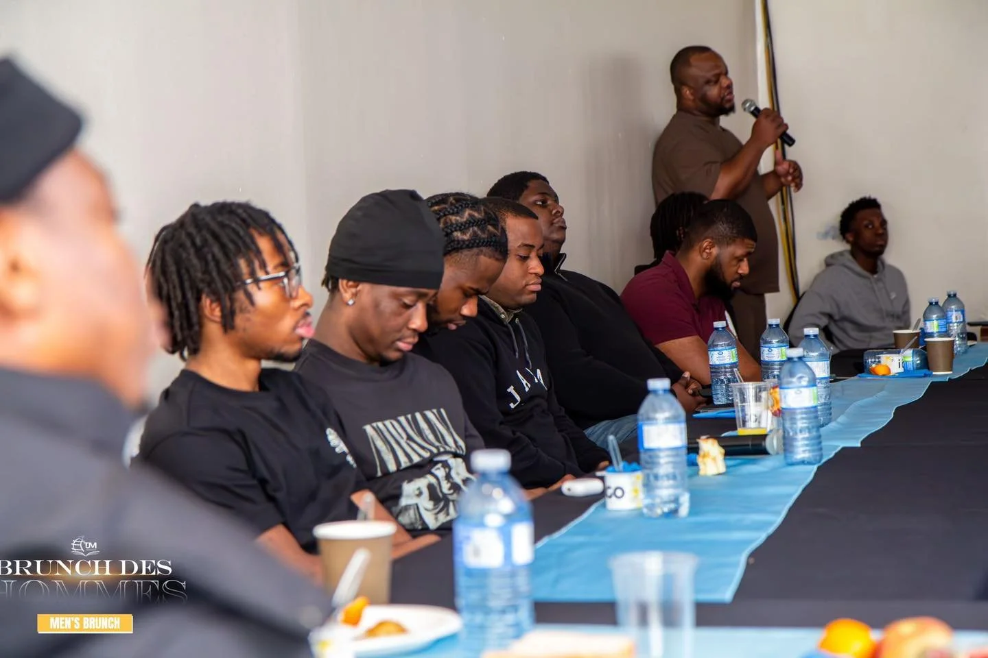 A group of men sitting at a long table during a brunch meeting, with water bottles and plates of food, while a man standing at the end speaks into a microphone.