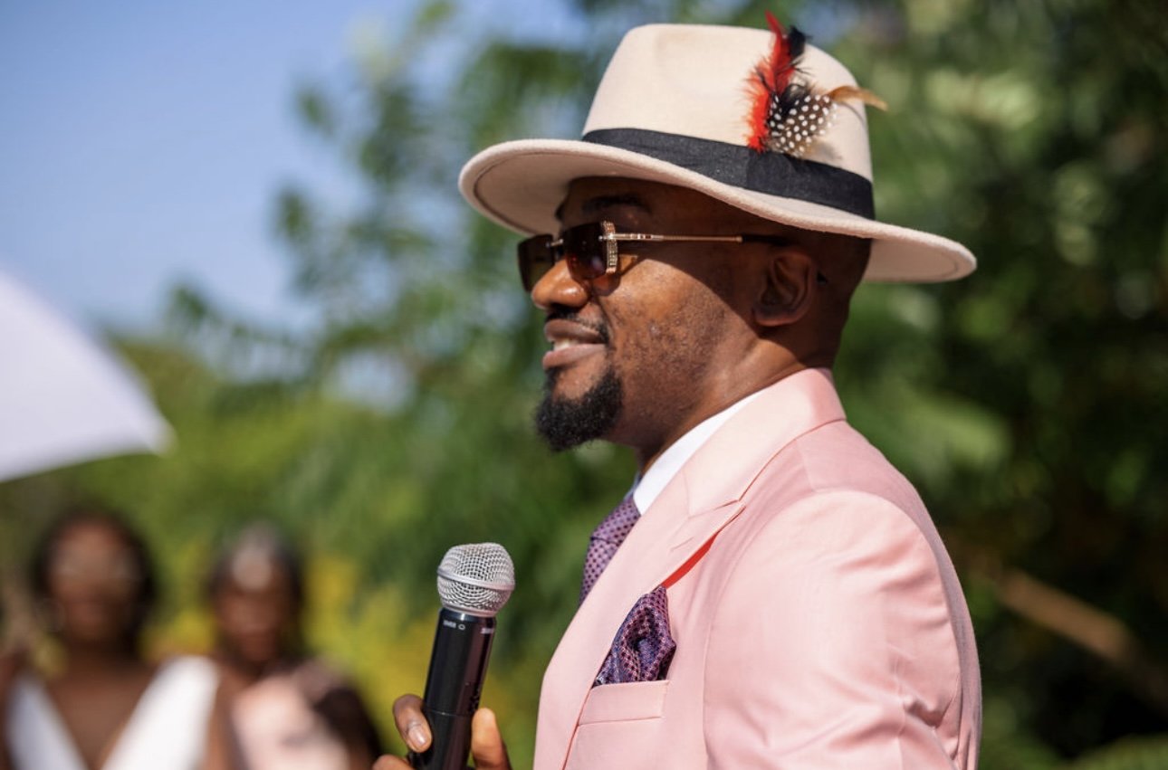 A man in a pink suit and a wide-brimmed hat with feathers and a feathered decoration, wearing sunglasses, speaking into a microphone outdoors during daytime, with blurred women in the background.