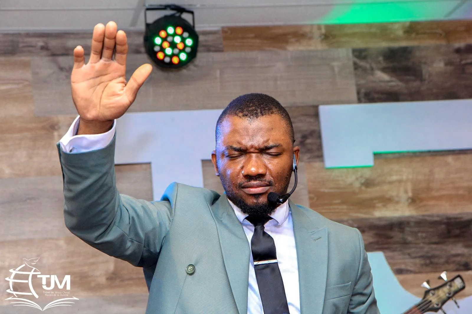 A man dressed in a suit with a tie, eyes closed, raising his right hand in prayer or worship, wearing a headset, standing in front of a wooden wall with a colorful stage light above him.