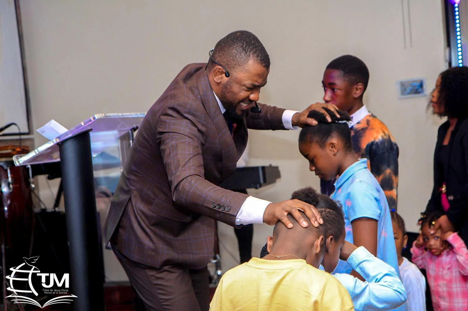 A man in a brown suit is praying over children with his hands on their heads, in a church setting with a lectern and keyboard in the background, and other children and adults watching.