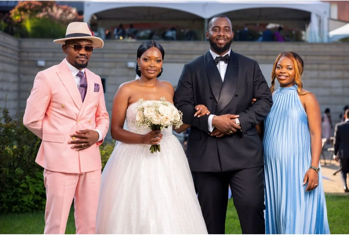 Group of four people dressed in formal attire at an outdoor wedding or special event, with a woman in a white wedding gown holding a bouquet of white roses and three men and women in colorful suits and dresses.