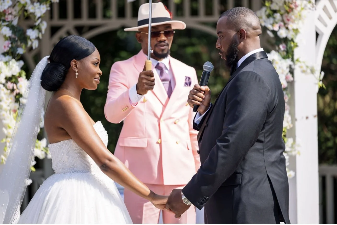 Bride and groom holding hands during their wedding ceremony outdoors, with a man in a pink suit and hat officiating.
