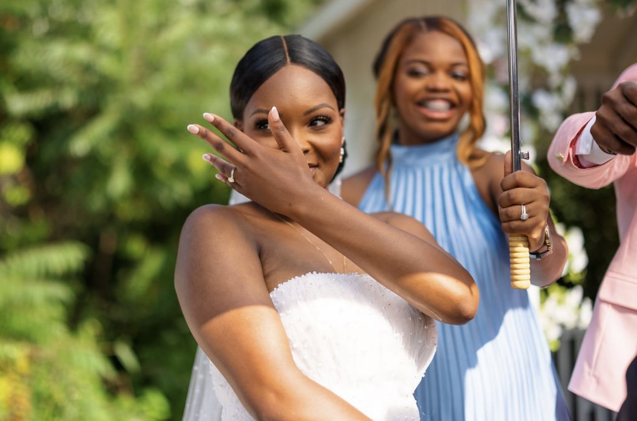 A bride in a white wedding dress shows her engagement ring to the camera, smiling and surrounded by lush green foliage. A woman in a blue dress stands behind her, smiling and holding an umbrella.