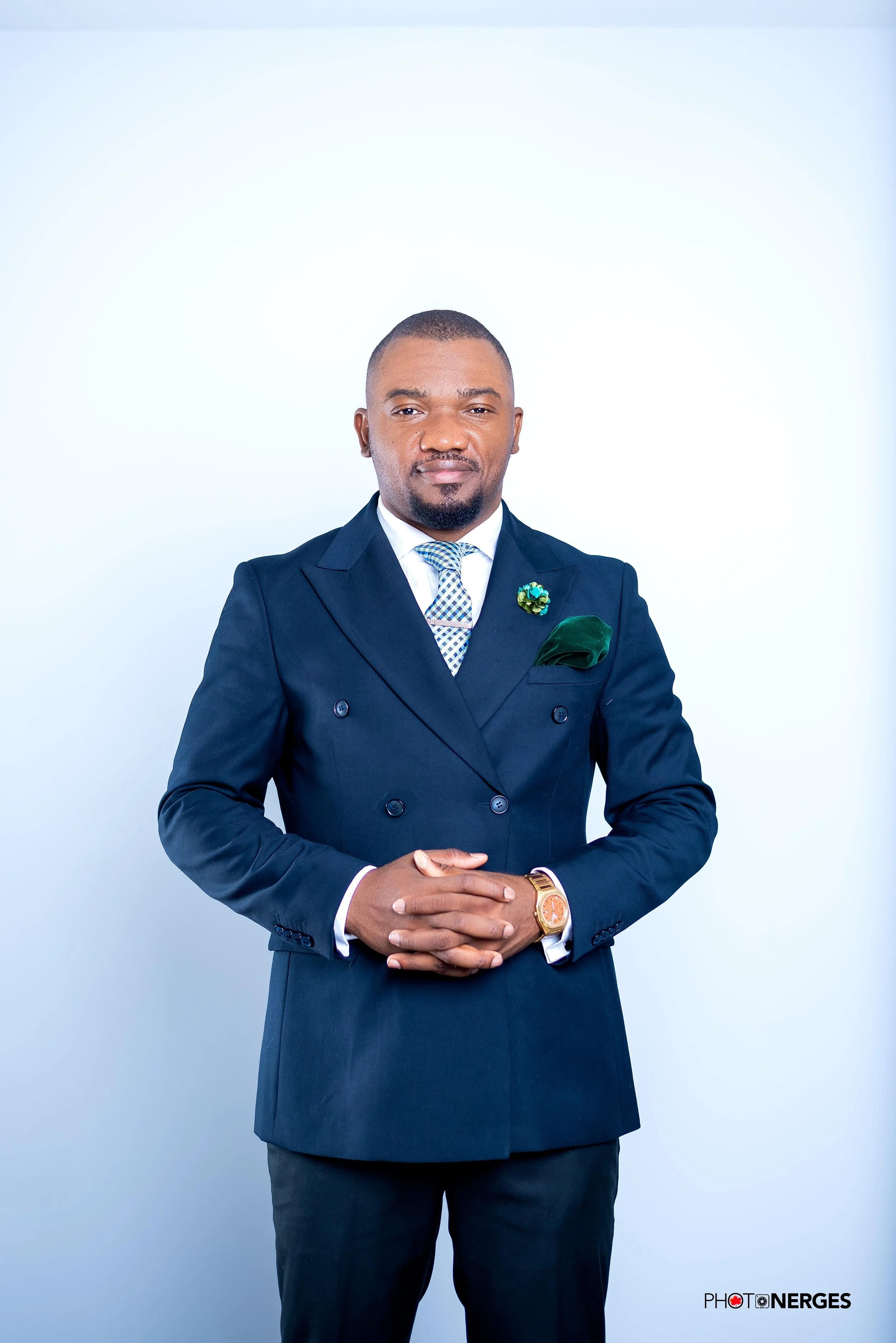 A man in a dark blue double-breasted suit with a patterned tie and a green pocket square, standing with hands clasped in front of a plain light background.