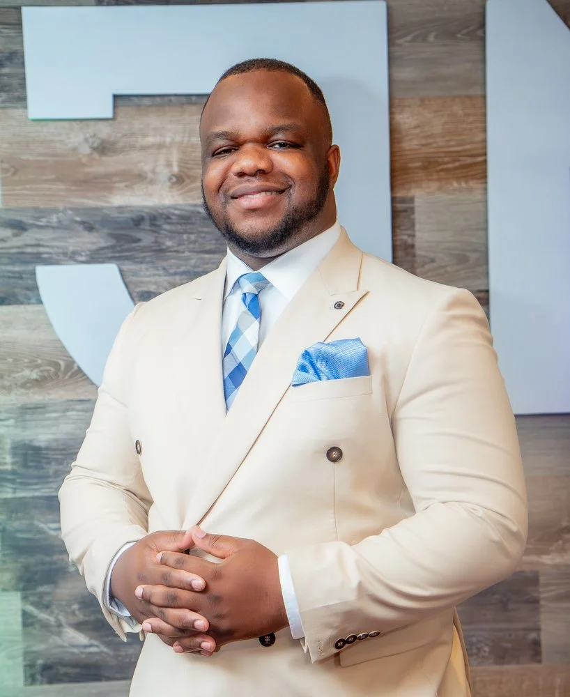 A confident man in a light beige suit with a blue plaid tie and matching pocket square, standing with hands clasped in front of a modern wooden wall with a light blue and white logo or artwork behind him.