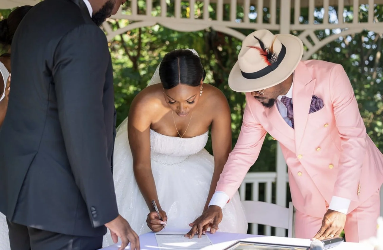 Bride signing a marriage certificate at her wedding, with a woman in a pink suit and a man in a black suit observing, outdoors with greenery in the background.