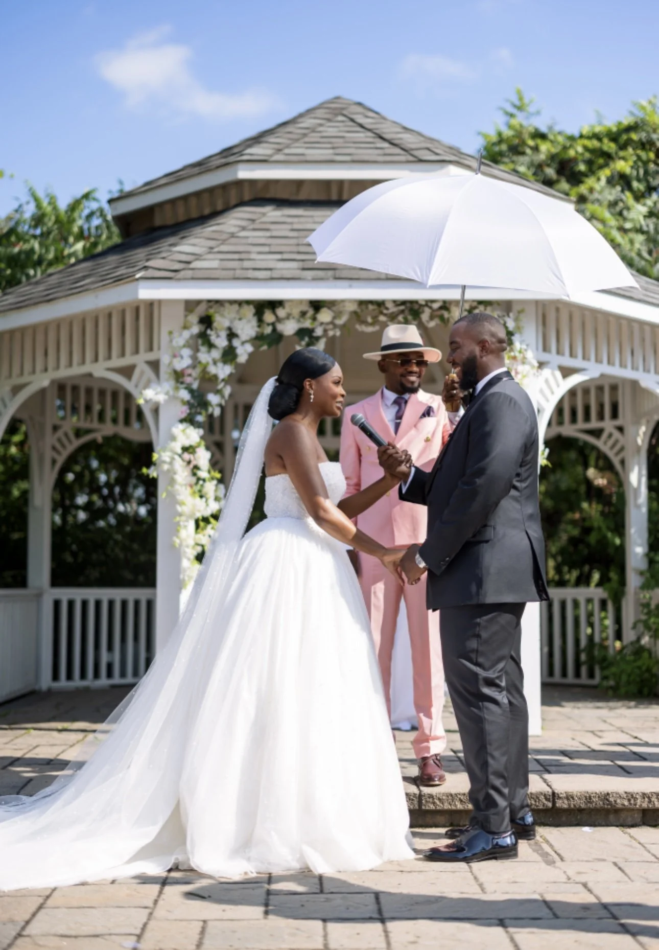 A wedding ceremony with a bride and groom holding hands and smiling, standing under a white umbrella, with an officiant in pink attire behind them in front of a decorative gazebo on a sunny day.