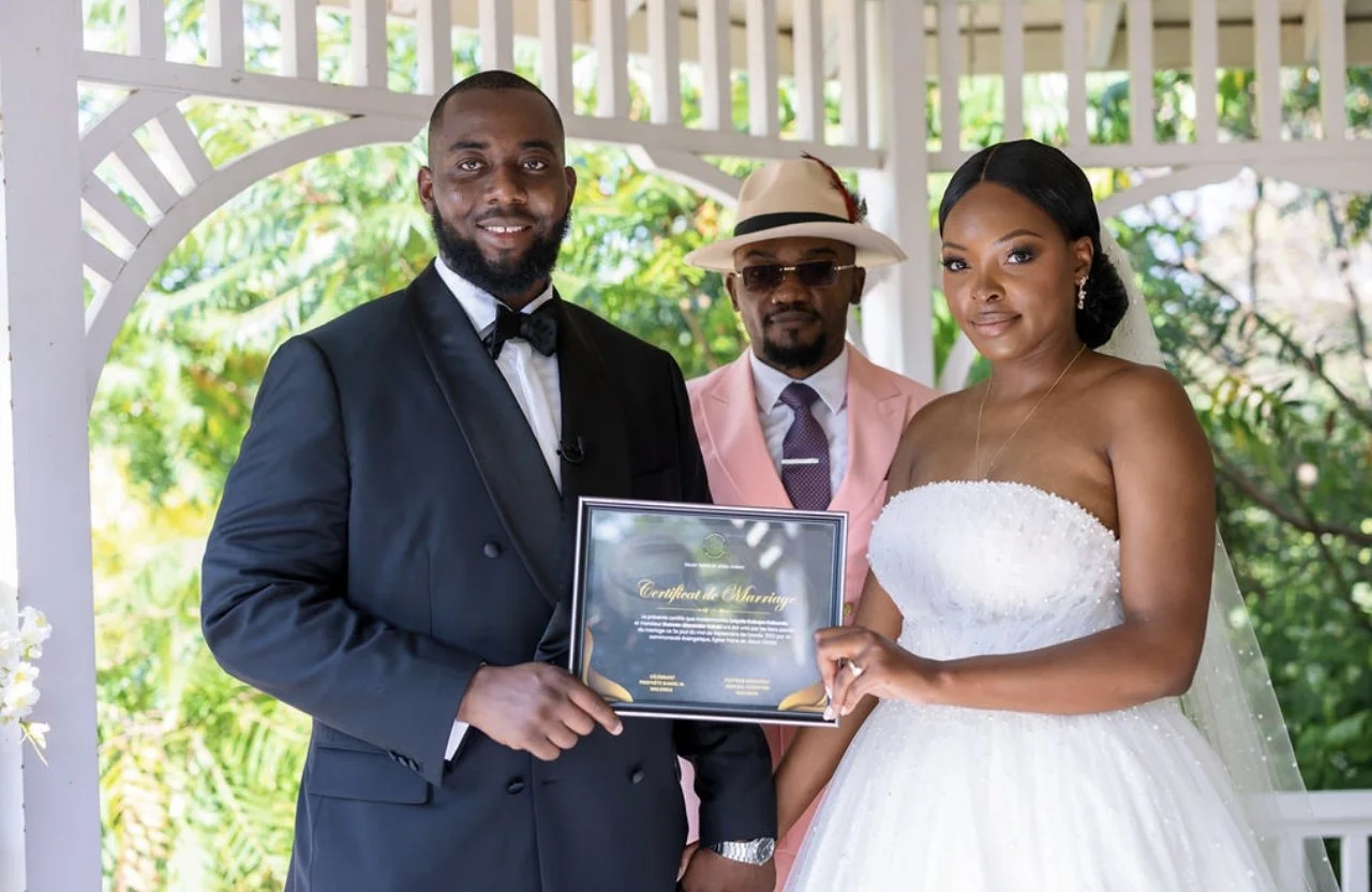 A bride and groom holding a marriage certificate, standing with an officiant outdoors during a wedding ceremony