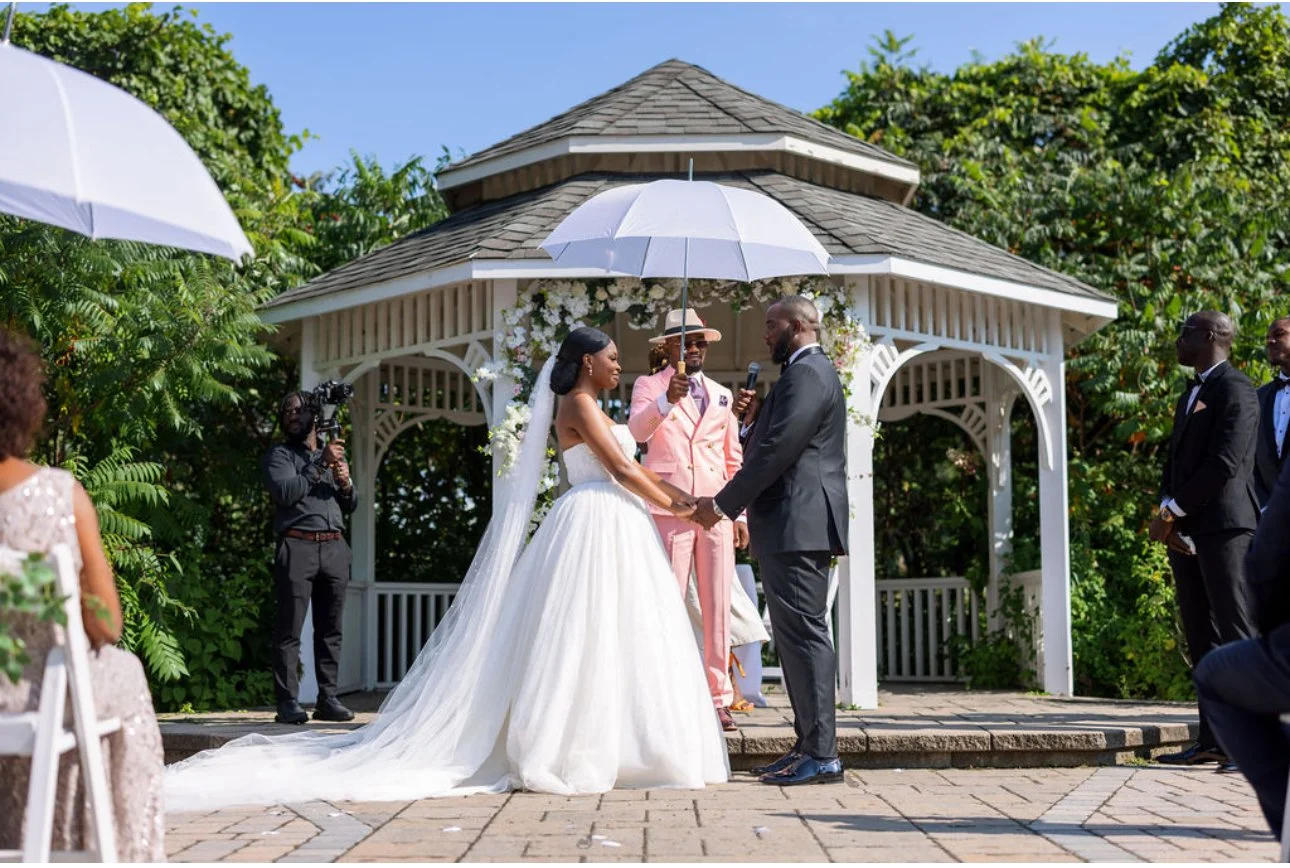 A wedding ceremony taking place outdoors under a small white gazebo surrounded by greenery, with the bride and groom holding hands and exchanging vows. The bride wears a white wedding gown and veil, while the groom is in a black suit. An officiant in