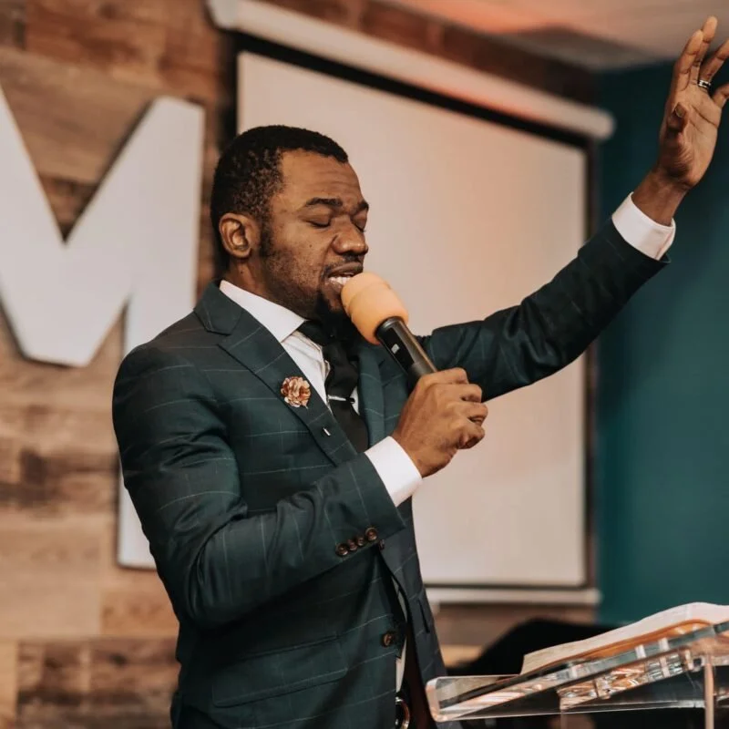 A man in a suit holding a microphone and raising his other hand, speaking passionately during a sermon in a room with a wooden wall and a large screen behind him.