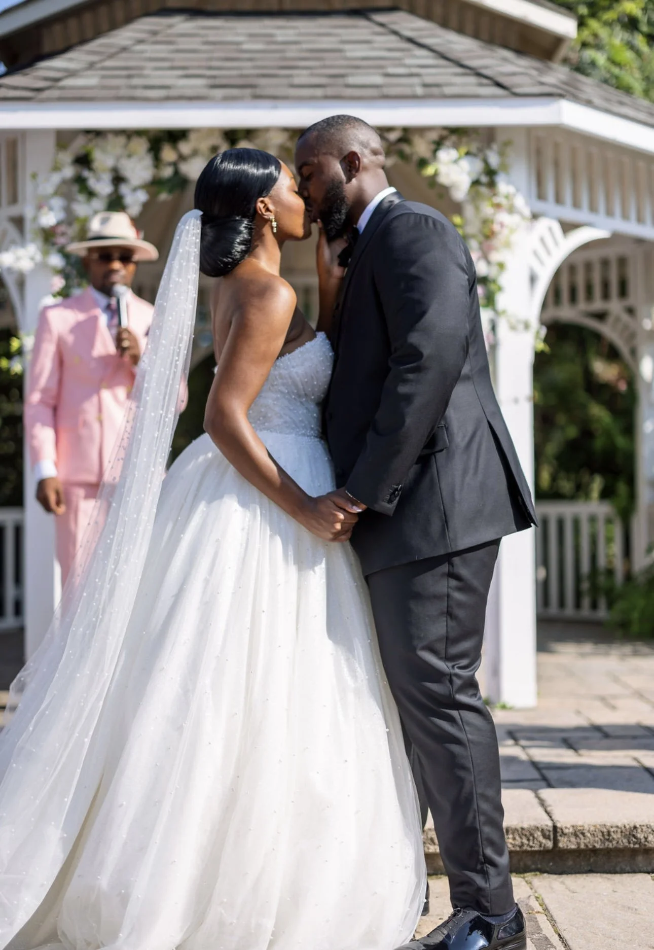 A bride and groom kiss during their wedding ceremony outdoors under a decorated pavilion, with an officiant in the background.