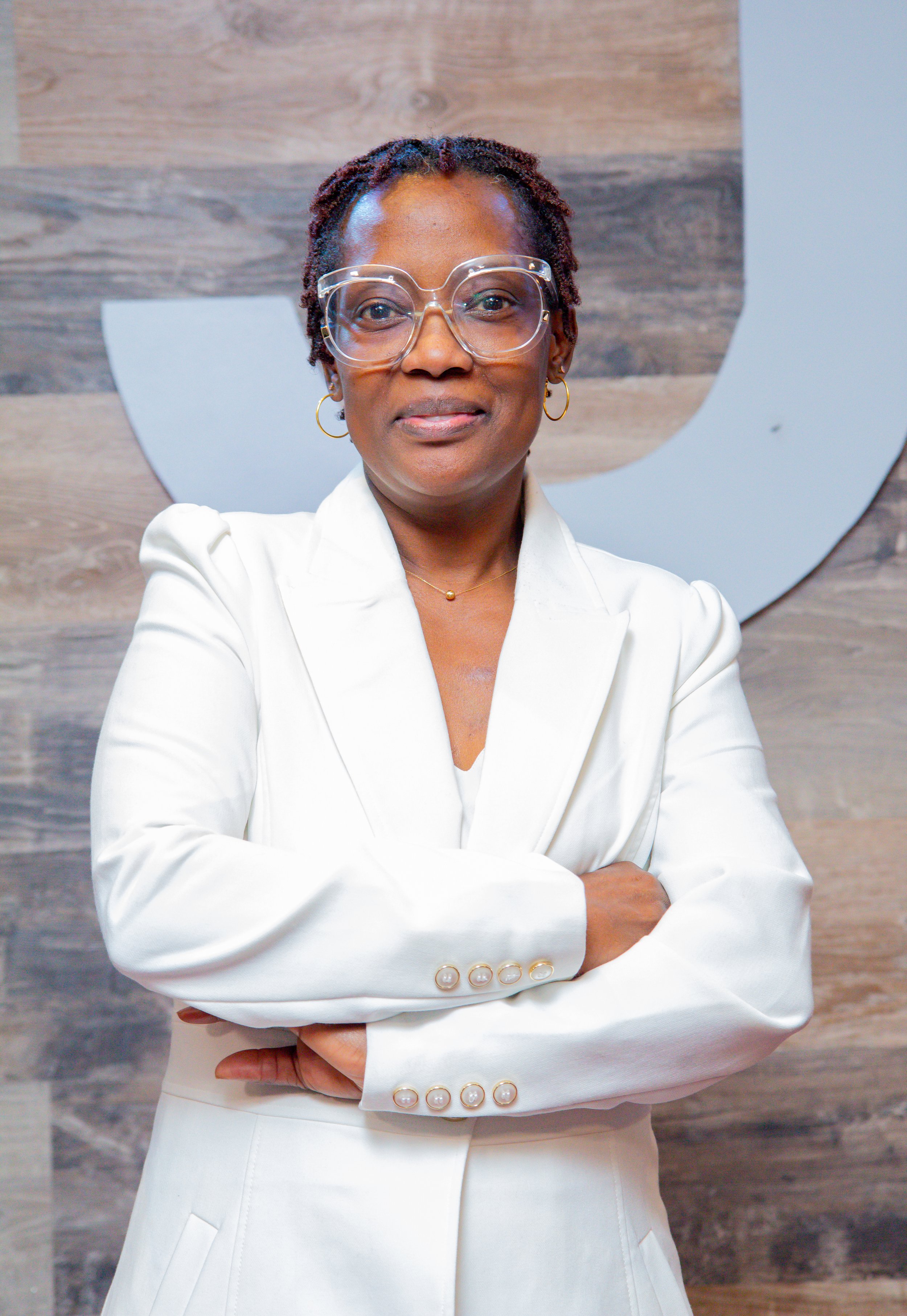 A woman with short braided hair wearing clear glasses and a white blazer standing with her arms crossed in front of a wall with a company logo.