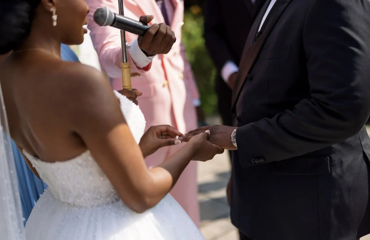 A woman in a white wedding dress is holding a man's hand during a wedding ceremony, with a person in a pink suit holding a microphone nearby.