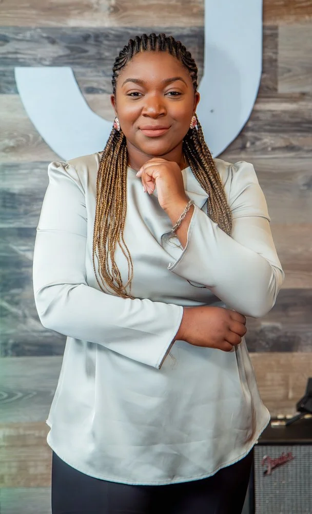 A woman with braided hair wearing a shiny cream-colored blouse, standing in front of a wooden wall with a large white letter 'U' and a small amplifier on the floor.