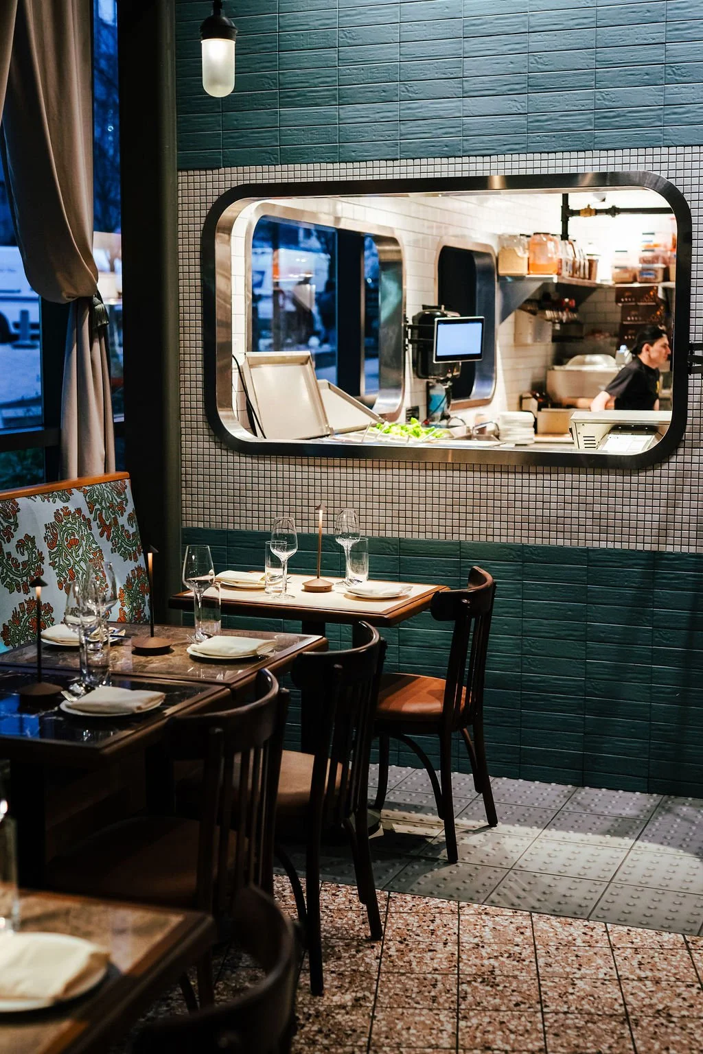 Empty dining table with glasses and napkins in a restaurant, view through a window into the kitchen with a staff member working.