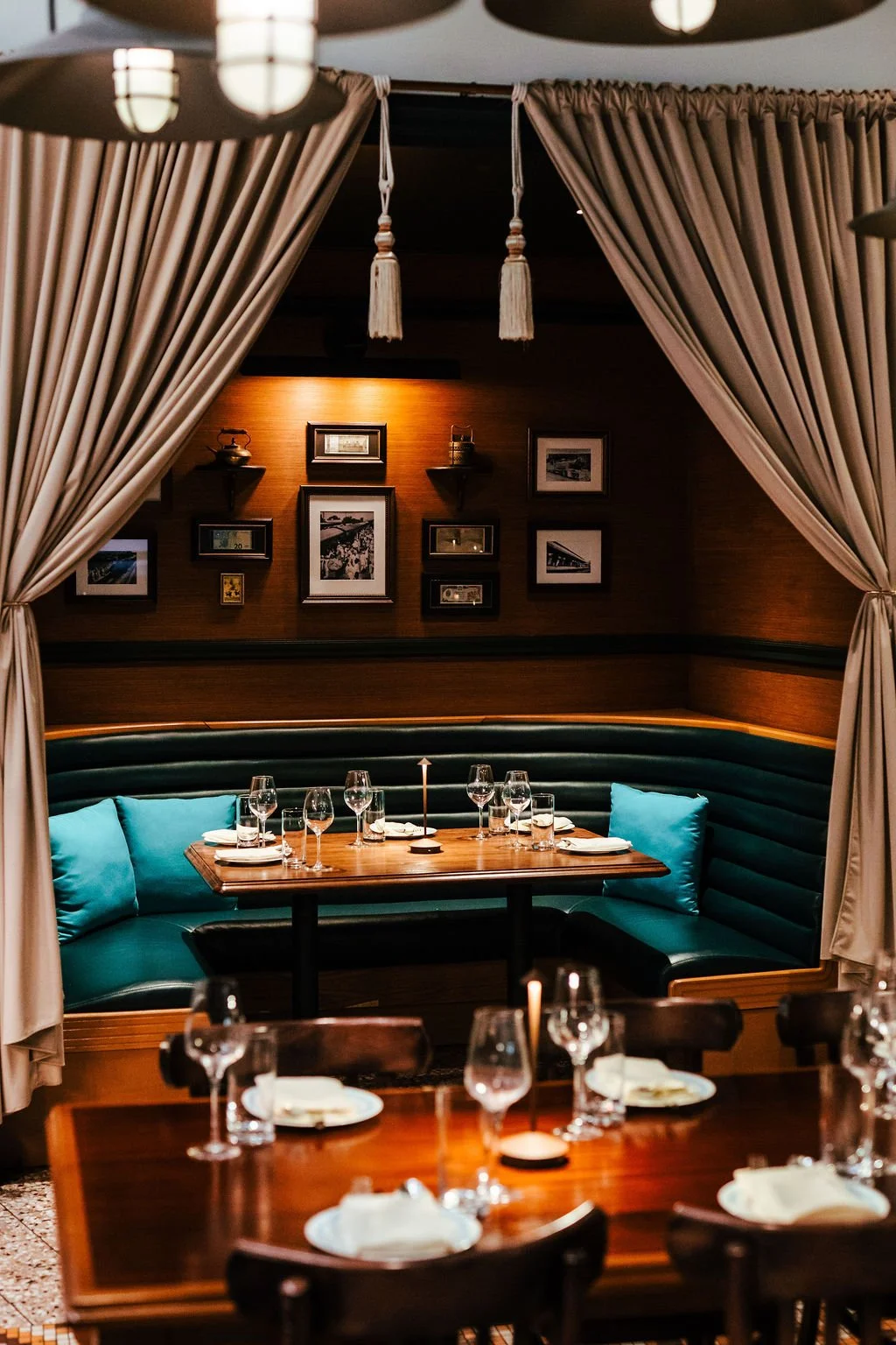 A cozy restaurant corner with a wooden booth featuring teal cushions, set with wine glasses, plates, and napkins, framed black and white photographs, and beige curtains framing the scene.