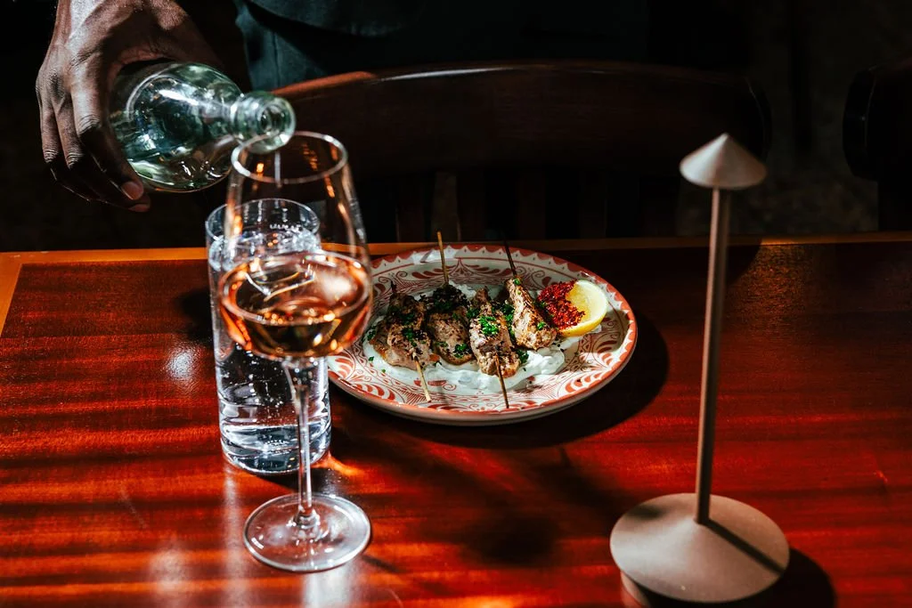 A table with a glass of rosé wine, a bottle of water, a plate of grilled skewered meat with a lemon wedge, and a modern table lamp.