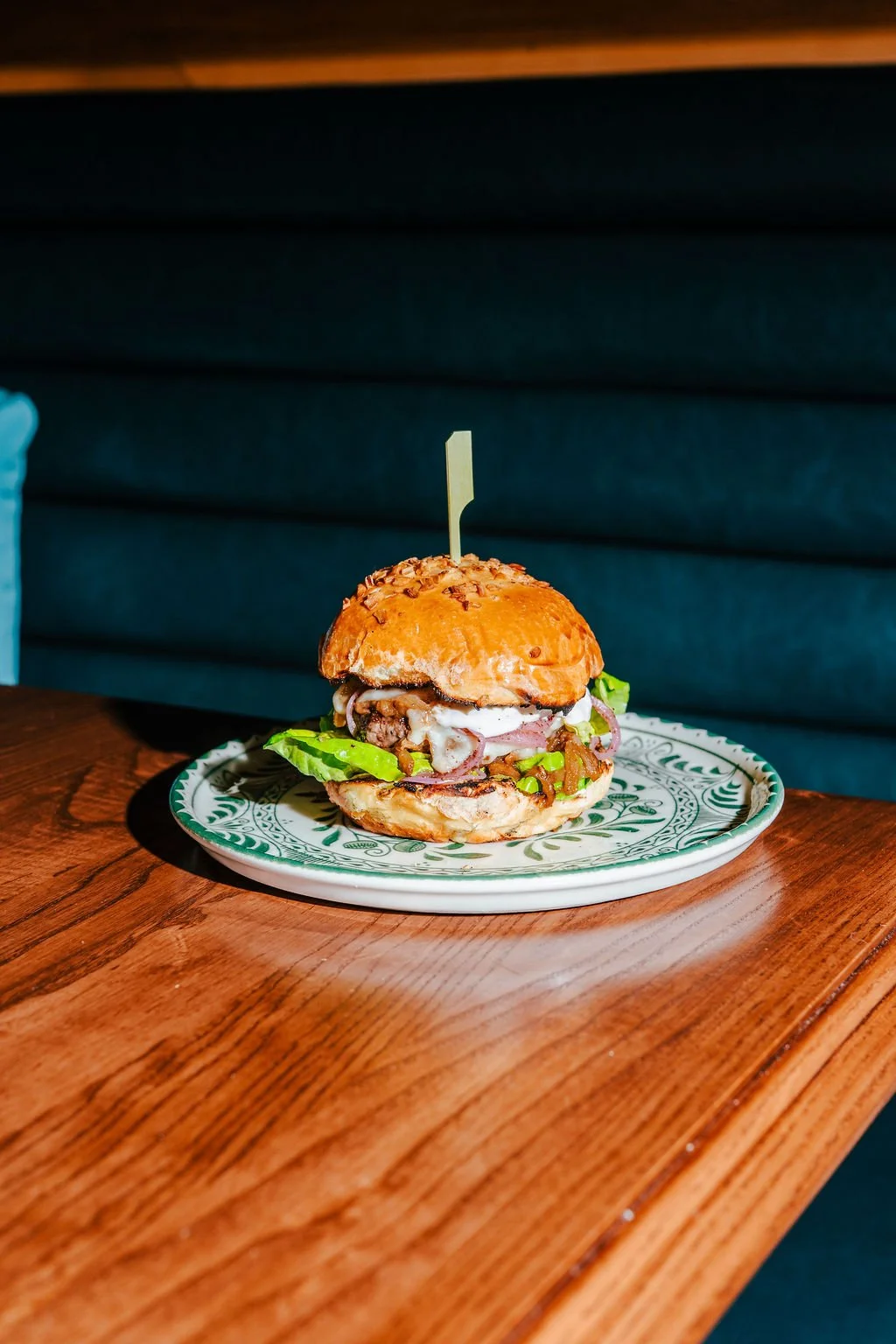 A cheeseburger with lettuce, pickles, onions, bacon, and sauces on a decorative plate, placed on a wooden table.