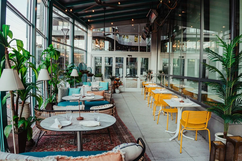 A bright, modern restaurant with floor-to-ceiling glass windows, green plants, white tables, yellow chairs, and a cozy seating area with blue cushions.