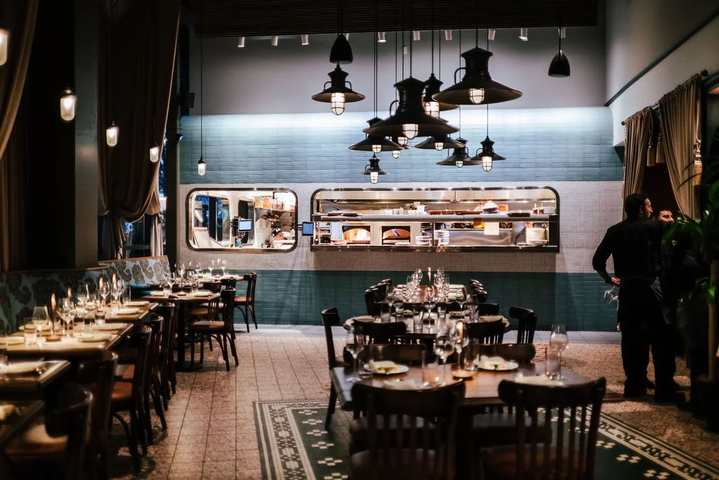 Interior of a restaurant dining area with tables set for a meal, overlooking an open kitchen through a large window, with hanging pendant lights and waitstaff preparing for service.