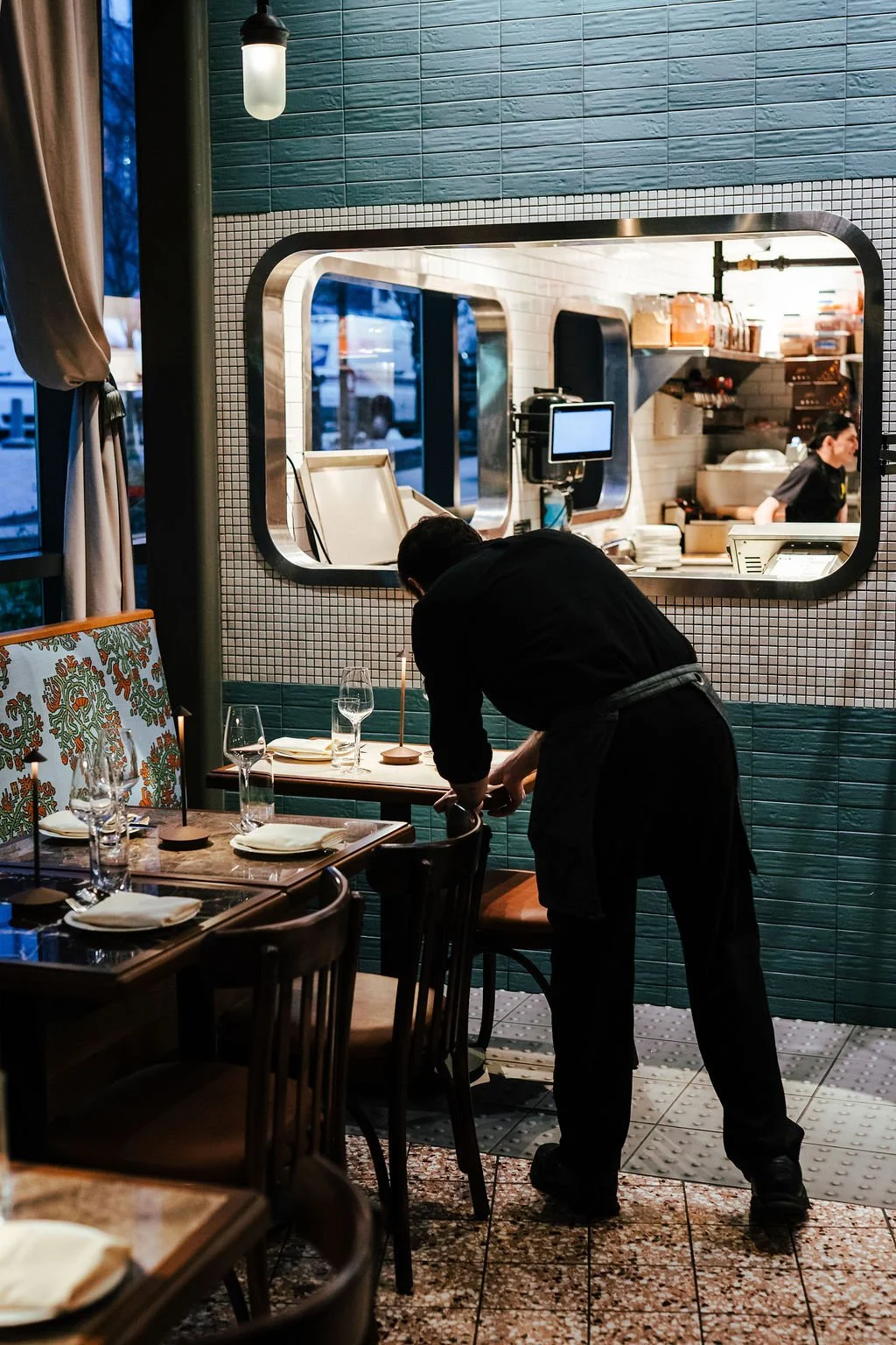 A waiter setting a table in a restaurant with a window to the kitchen visible.