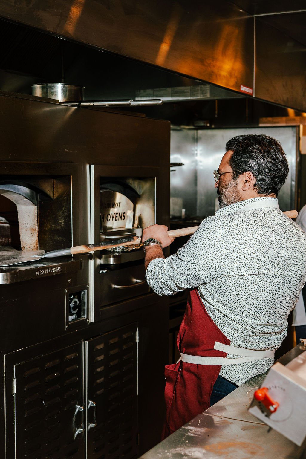 A man in glasses and a patterned shirt wearing a red apron bakes in a commercial oven.
