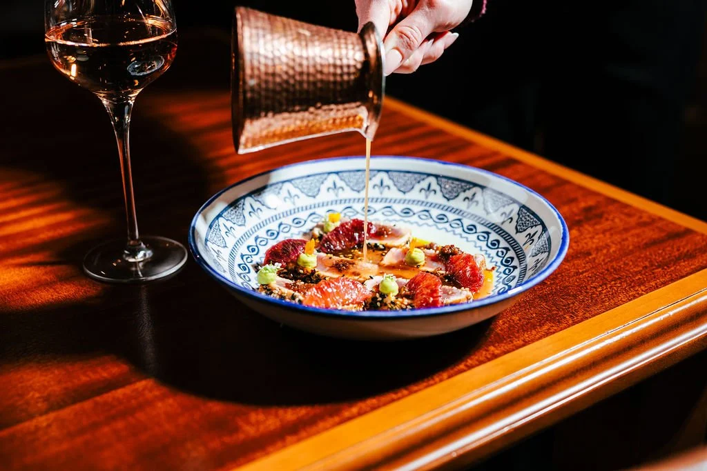 Person pouring sauce over a bowl of tuna carpaccio garnished with capers and herbs, next to a glass of rosé wine on a wooden table.
