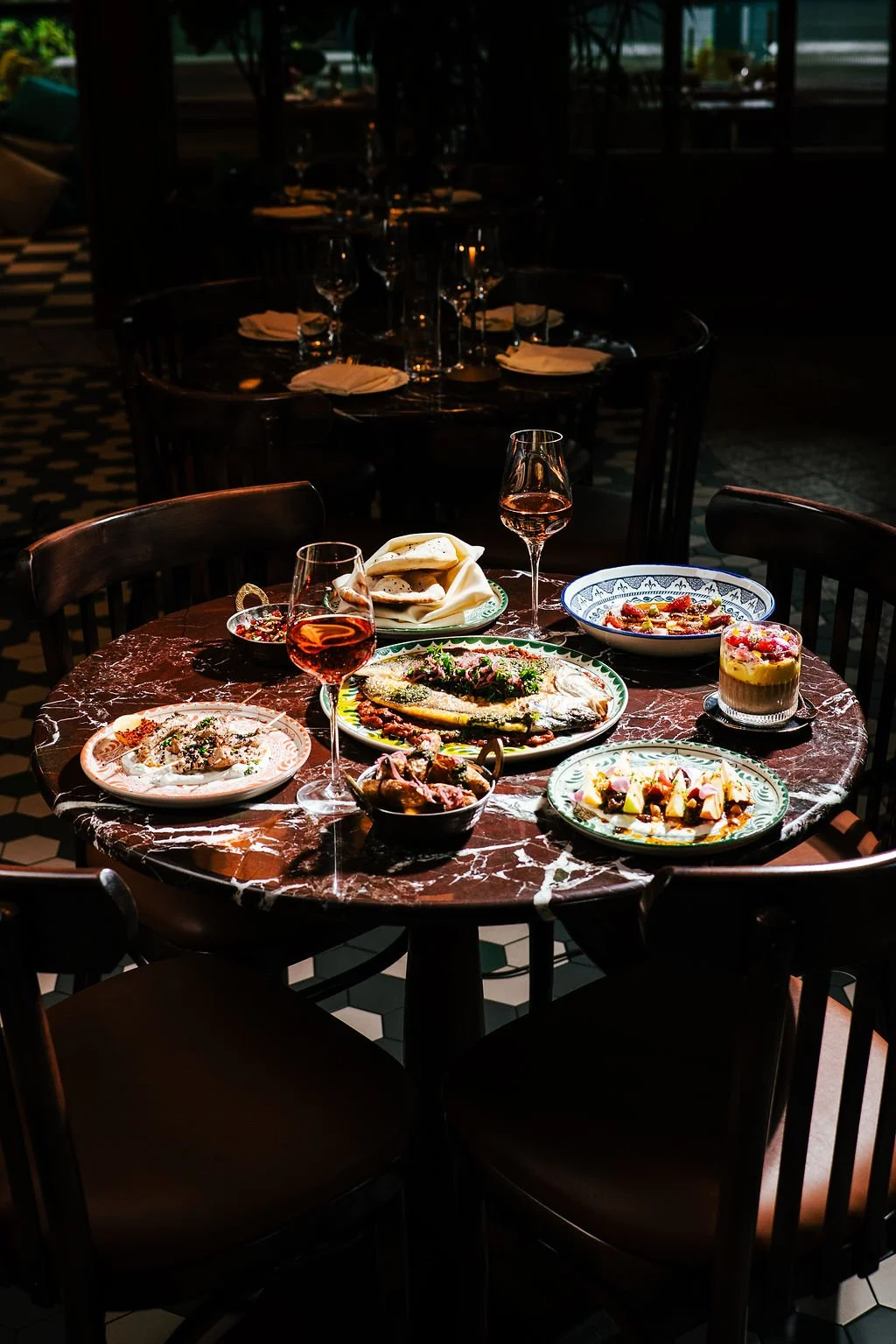 A round wooden table with a marble top set for dining, featuring various plates of food, two glasses of rosé wine, and bread in a dimly lit restaurant with a checkered floor and dark chairs.