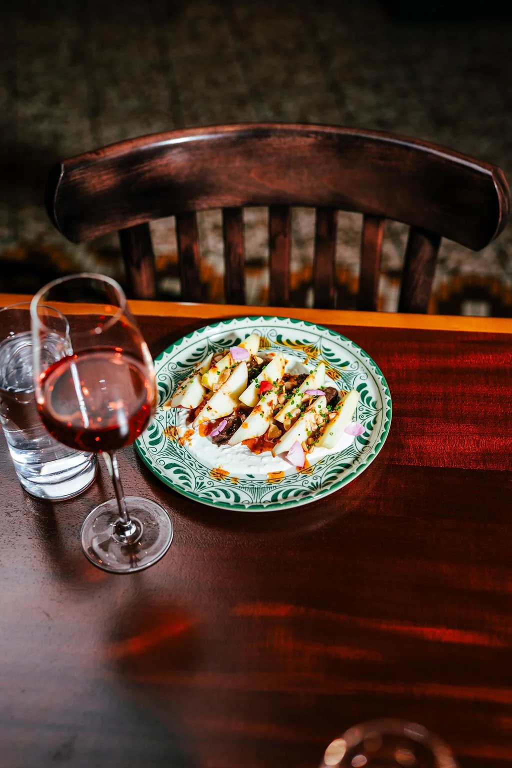 A dinner table with a plate of food, a glass of red wine, and a glass of water. The dish appears to be sliced cheese with herbs and garnishes on a decorative green and white plate.