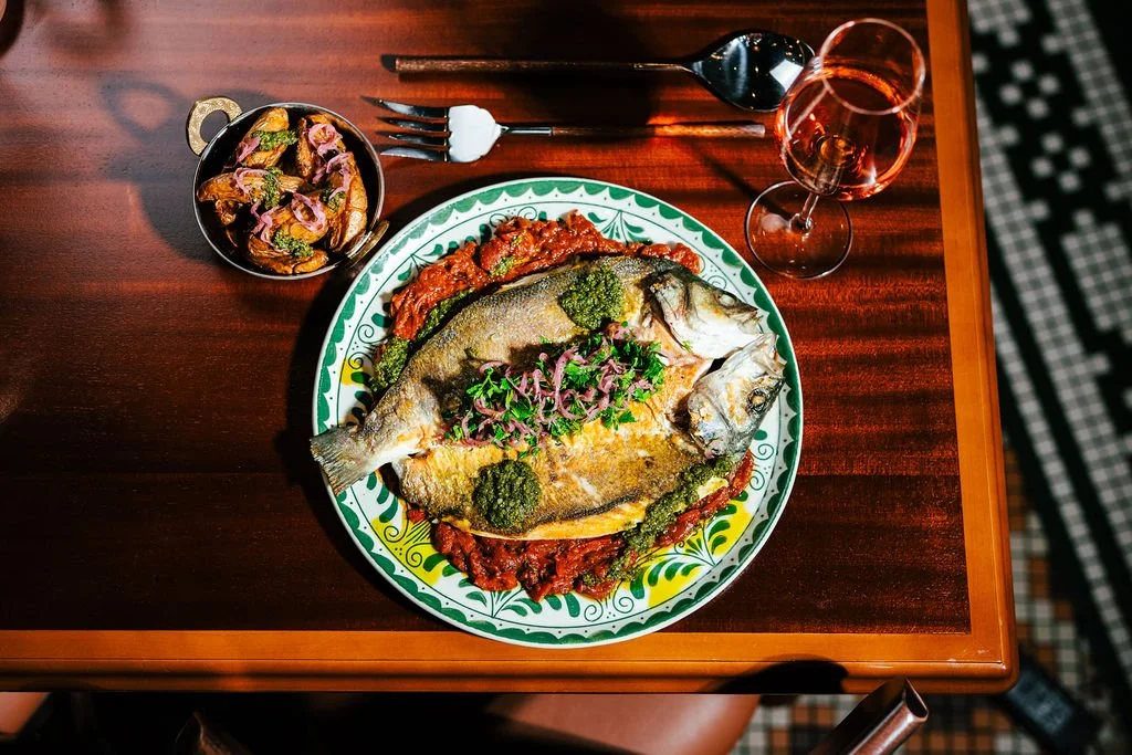 A plate with a cooked fish topped with greens and herbs, surrounded by red sauce and green sauce, on a decorative green and white plate. Next to it is a small bowl of cooked vegetables, a fork and knife, a glass of rosé wine, and a spoon on a wooden 