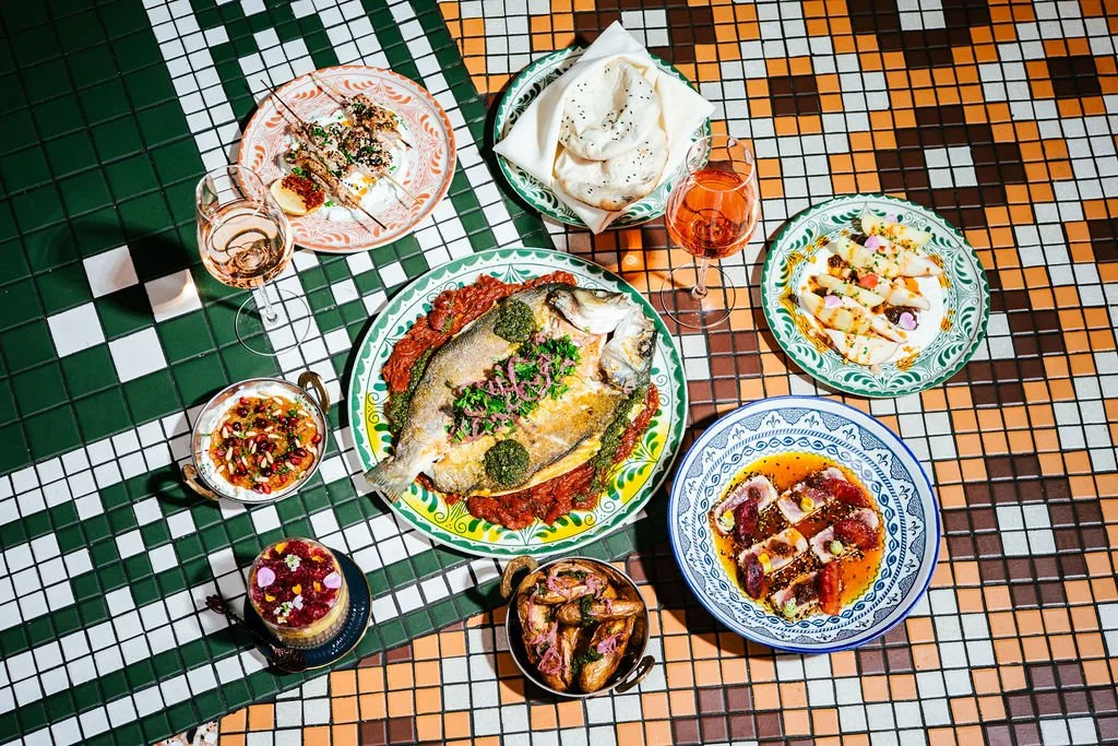 A colorful mosaic tiled table set with various dishes including a whole fish with sauce, a small bowl of sauce, a plate with sliced meat and sauce, a bowl of white bread, a glass of rosé wine, a bowl of chips, and a glass of rosé wine. The dishes are