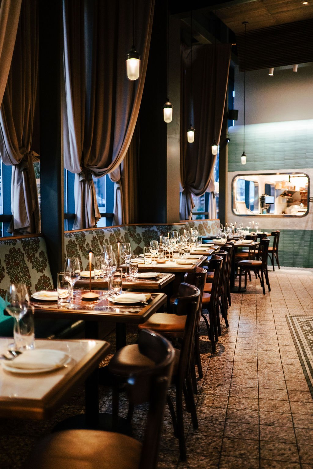 Empty dining tables with glassware and napkins in a restaurant with tall curtains and a window, dim lighting, and a view into the kitchen.