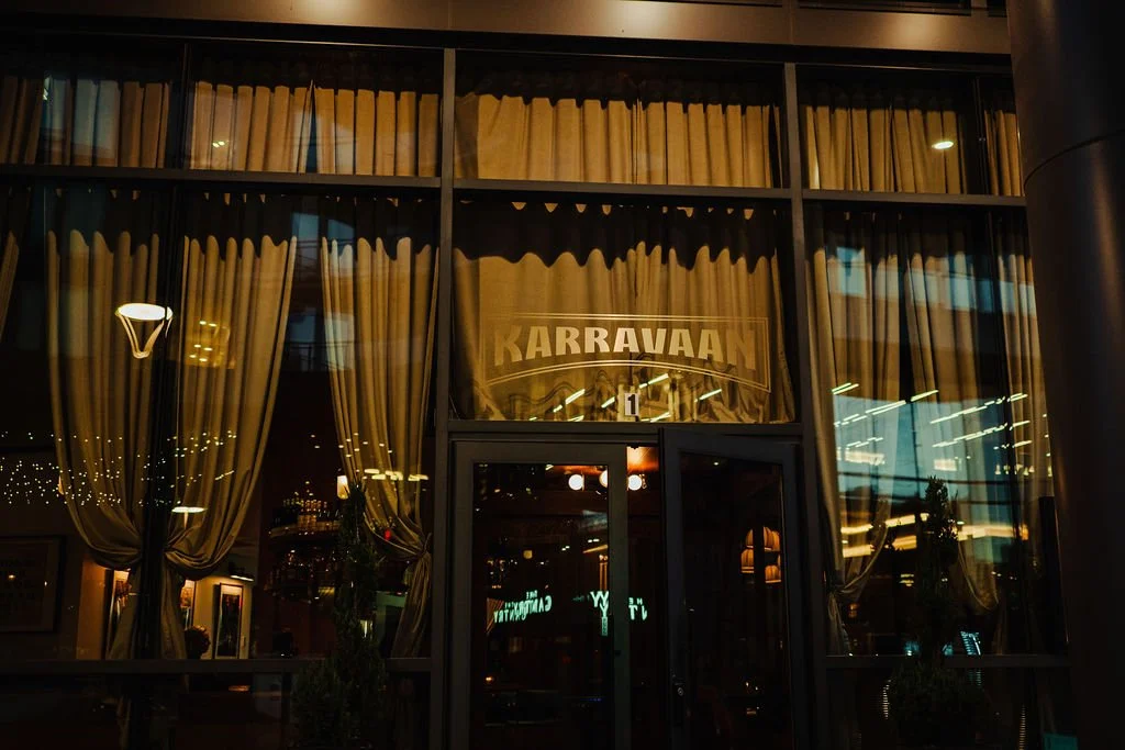 Night view of a restaurant or cafe named 'KARRAVAAN' with large window panes, beige curtains, and warm interior lighting.